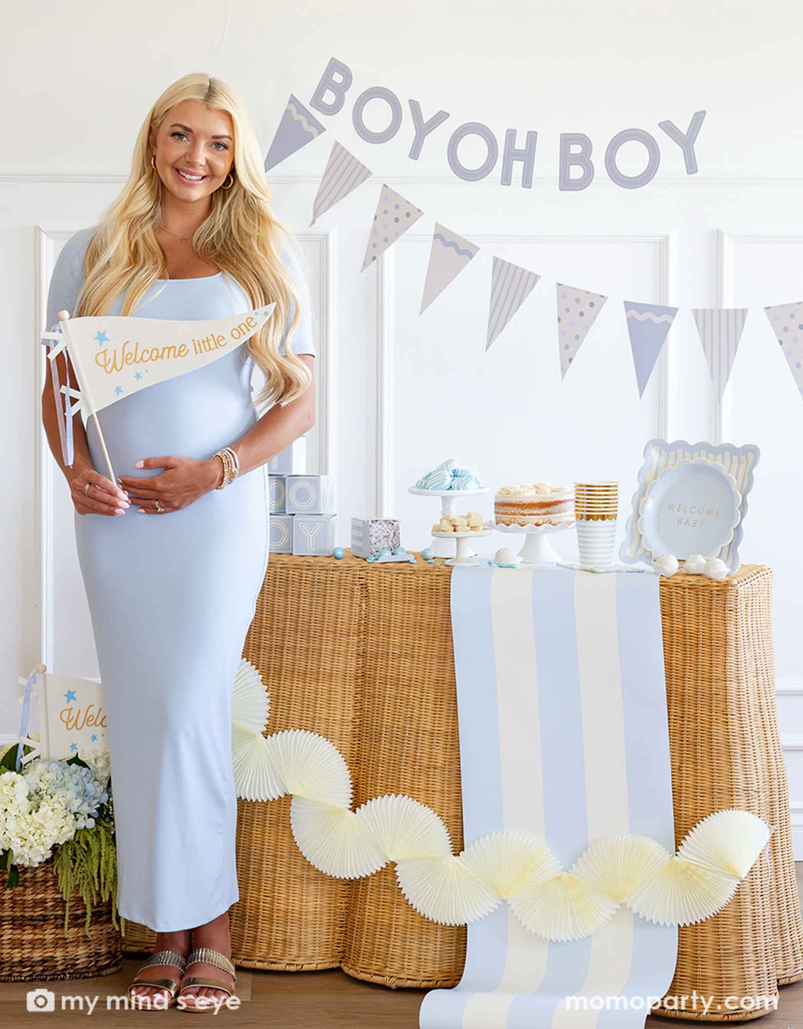 A modern and clean boy baby shower set up featuring various baby blue themed party supplies from Momopartys including party plates, cups, napkins, centerpieces on the table. A pregnant mom-to-be is standing in front of a baby shower setup with 'BOY OH BOY' banner and dessert table. She's holding a party pennant that says "Welcome to the World". With beautiful flower bouquet and cream colored bunting party banner around her.