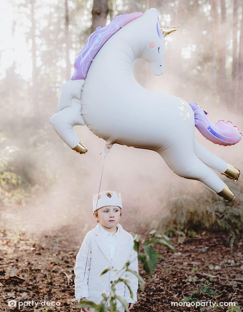 A boy dressed in white clothing with a party crown hat in an enchanting forest with Momopartys's enchanting unicorn shaped foil balloon floating in the air, making this a dreamy scene for kid's special celebration.