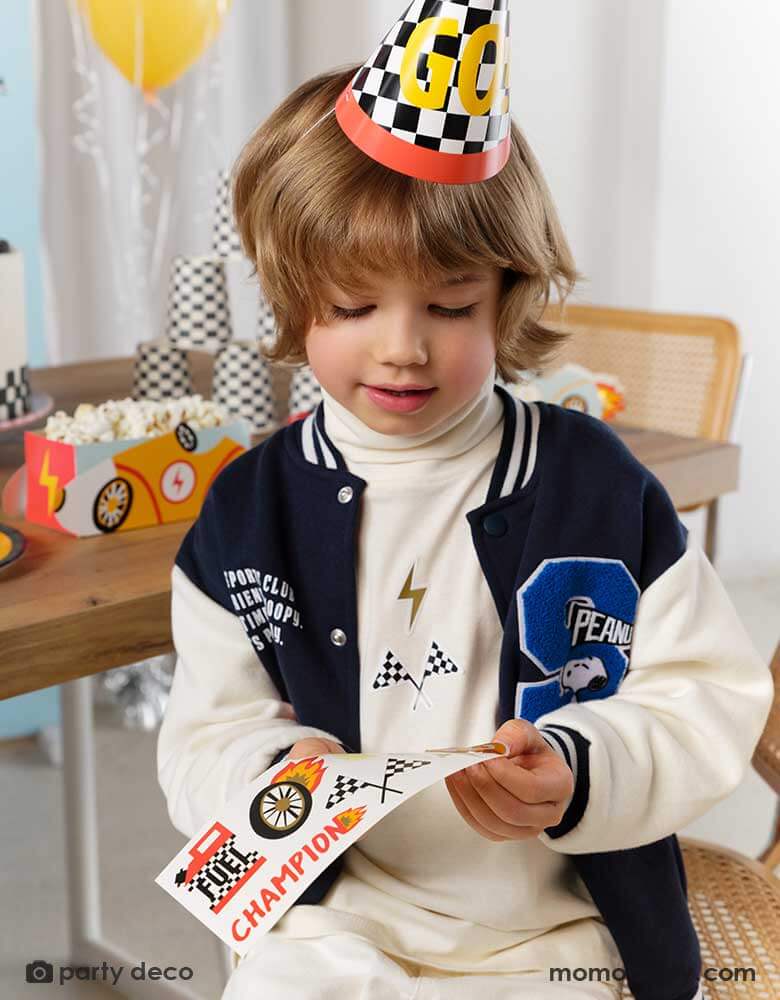 A little boy wearing a checkered flagged party hat with GO! on it in a race car party. In his back is a party table filled with race car themed treat boxes with popcorn in them. He's holding Momopartys's race car sticker sheets by Party Deco - a perfect party activity for boy's race car themed birthday celebrations.