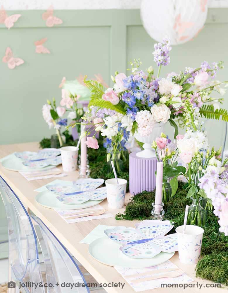 A beautiful butterfly themed party table featuring Momopartys's Flutter butterfly shaped plates paired with pastel mint dinner plates and napkins, next to it is butterfly themed party cups and napkins. In the middle there's moss grass textured table runner with pretty spring flower arrangement. On the wall there are butterfly shaped paper decorations attached on the wall, making this a perfect inspo for kid's butterfly fairy themed birthday or a gorgeous spring Easter celebration. 