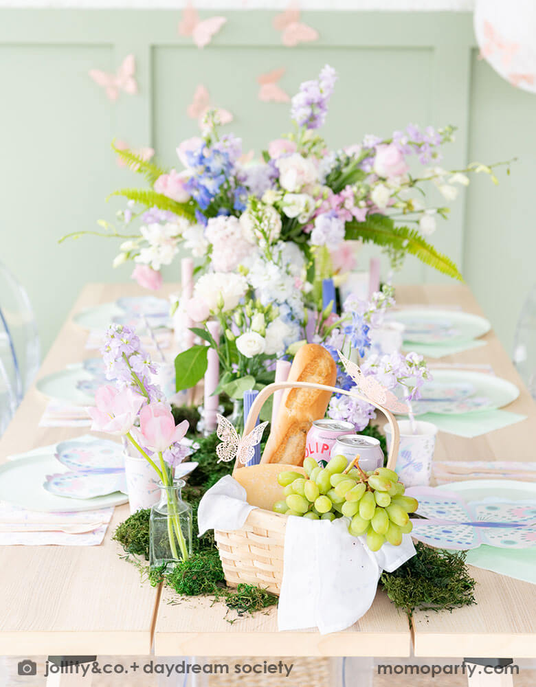 A beautiful butterfly themed party table featuring Momopartys's Flutter butterfly shaped plates paired with pastel mint dinner plates and napkins, next to it is butterfly themed party cups and napkins. In the middle there's moss grass textured table runner with pretty spring flower arrangement and a picnic basket of fruits and breads. On the wall there are butterfly shaped paper decorations attached on the wall, making this a perfect inspo for kid's butterfly fairy themed birthday or a spring celebration. 