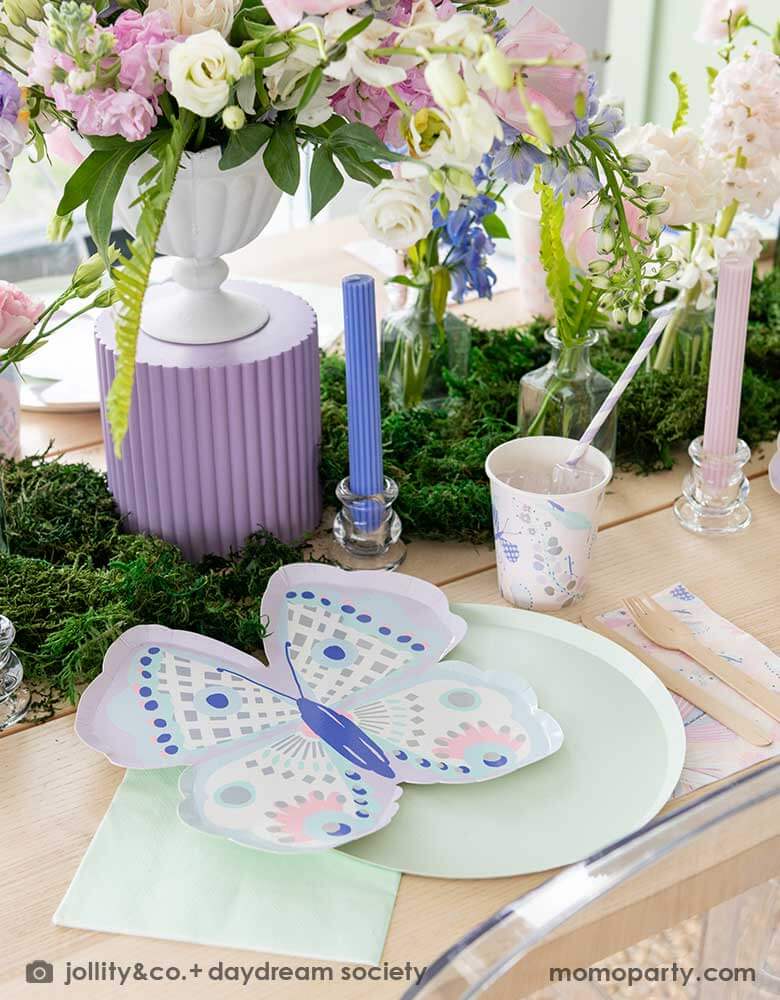 A beautiful butterfly themed party table featuring Momopartys's Flutter butterfly shaped plates paired with pastel mint dinner plates and napkins, next to it is butterfly themed party cups and napkins. In the middle there's moss grass textured table runner with pretty spring flower arrangement, making this a perfect inspo for kid's butterfly fairy themed birthday or a gorgeous spring Easter celebration. 