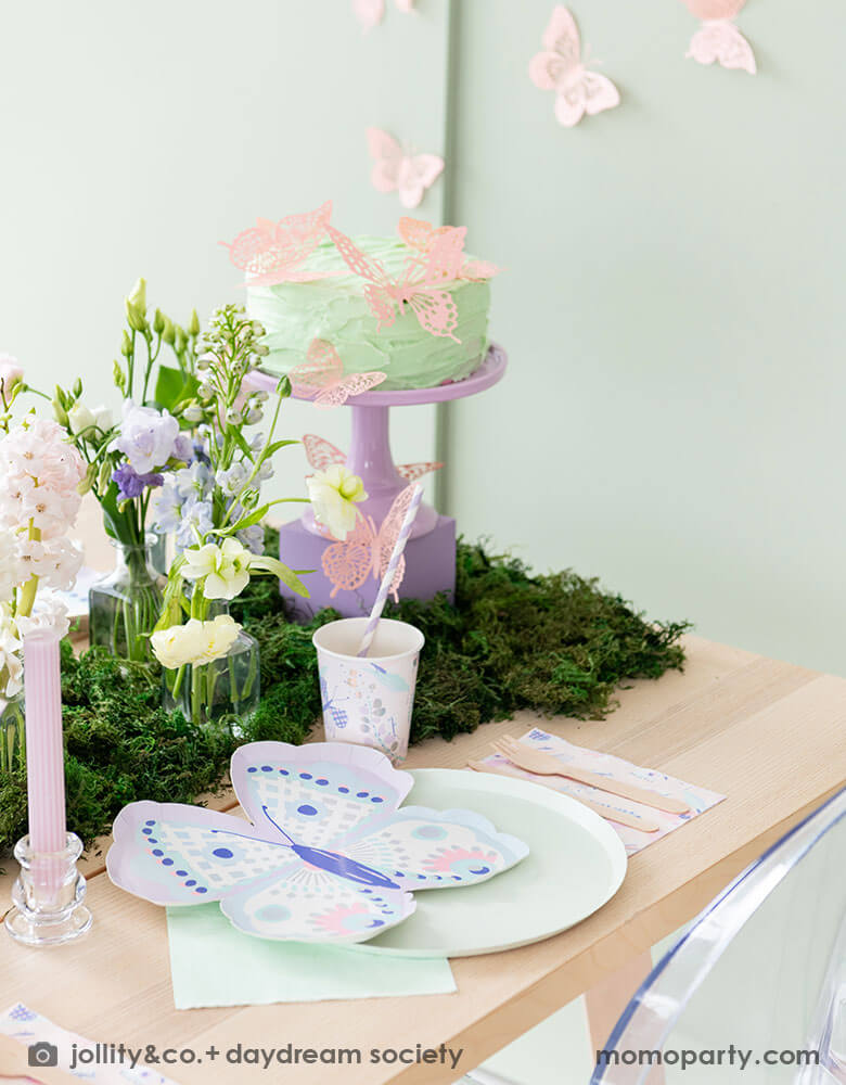 A beautiful butterfly themed party table featuring Momopartys's Flutter butterfly shaped plates paired with pastel mint dinner plates and napkins, next to it is butterfly themed party cups and napkins. In the middle there's moss grass textured table runner with pretty spring flower arrangement and paste pink candles. On the wall there are butterfly shaped paper decorations attached on the wall, making this a perfect inspo for kid's butterfly fairy themed birthday or a gorgeous spring Easter celebration. 