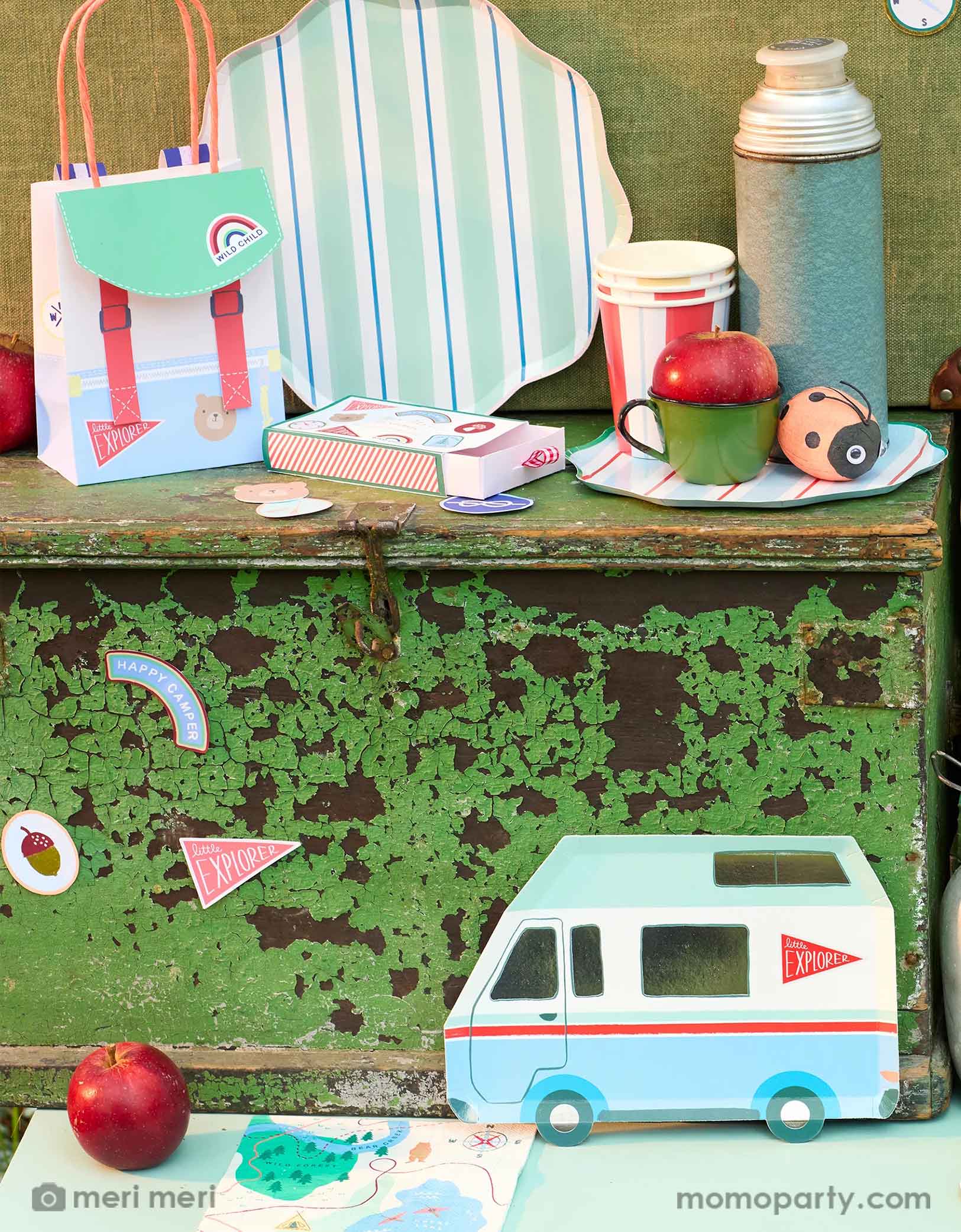 A camping themed birthday party display showcasing Momopartys's camping themed party supplies by Meri Meri including the camping RV van shaped plate, the camping maps napkins, the camping themed stickers, and camping backpack shaped party bag, along with striped dinner plates, party cups and a vintage thermos bottle with a ladybug surprise ball next to it, this camping adventure party collection is perfect for your nature-loving child's camping themed celebration.