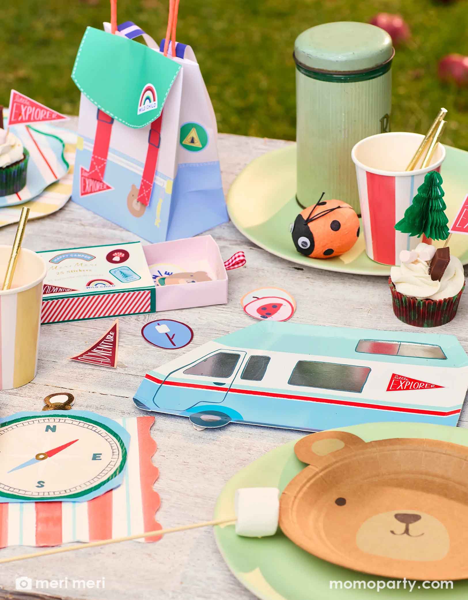 A camping themed birthday party table featuring Momopartys's camping themed party supplies by Meri Meri including the camping RV van shaped plate, the camping compass shaped napkins, the camping themed stickers, and camping backpack shaped party bag, along with striped dinner plates, party cups and a vintage thermos bottle with a ladybug surprise ball next to it, this camping adventure party collection is perfect for your nature-loving child's camping themed celebration.