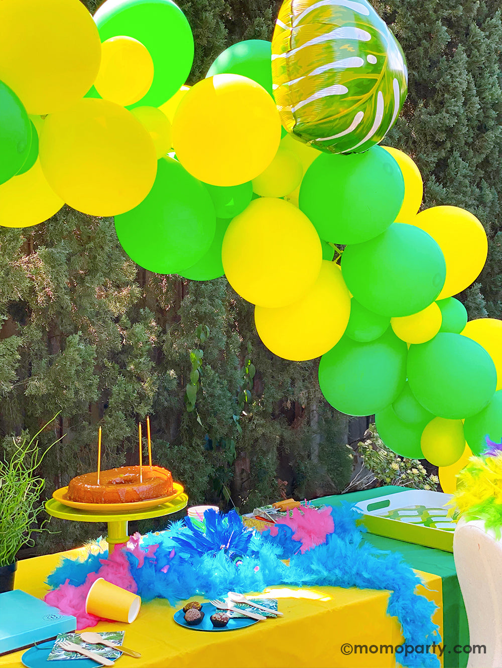 A festive green and yellow balloon arch decorated with Momopartys's 21" Palm Leaf Foil Balloon. On the table there are some Brazilian carnival themed feather decorations and a birthday cake. A perfect idea for a tropical, luau or island themed party in the summer - be it a summer backyard BBQ gathering, a birthday party or just a summer cookout event!