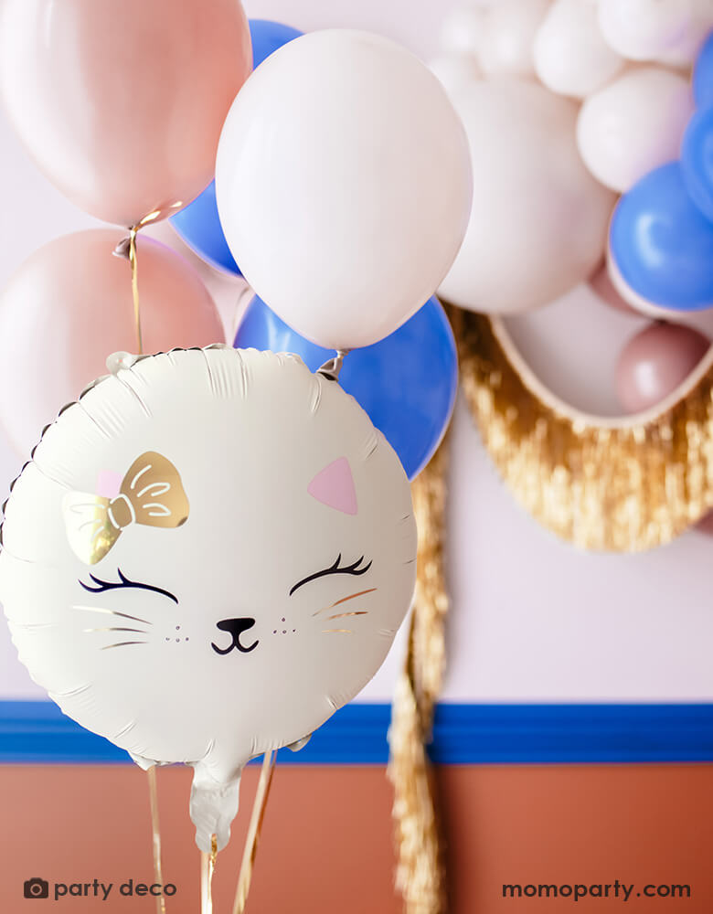 A cat themed party decoration set up features Momopartys's 18" kitty cat shaped foil balloon in cream, along with some latex balloons in pink, blush and royal blue adorned with gold tinsel garland hung on the wall, making this an adorable inspo for kid's kitty cat or pet themed party decoration.