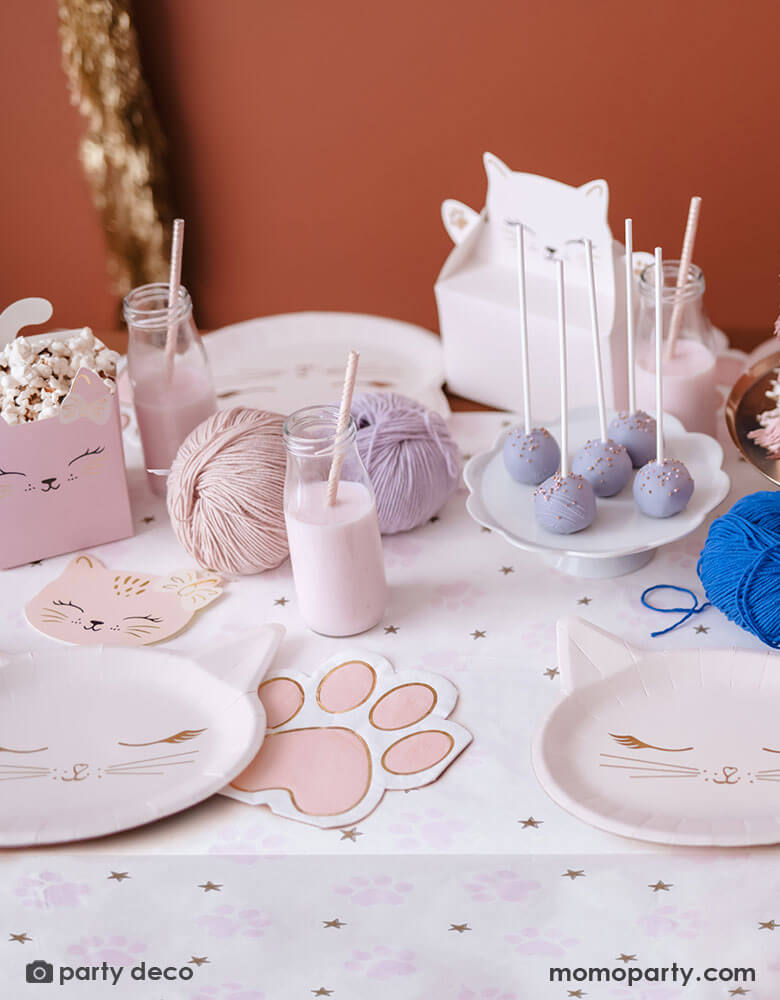 A cat themed party table featuring various cat themed party supplies from Momopartys including party plates, pink paw shaped napkins, cat shaped treat boxes filled with popcorn, small milk bottles with pale pink drink and a table of treats and snacks in pastel pink and lilac colors, making this an adorable inspo for kid's kitty cat themed birthday celebration.