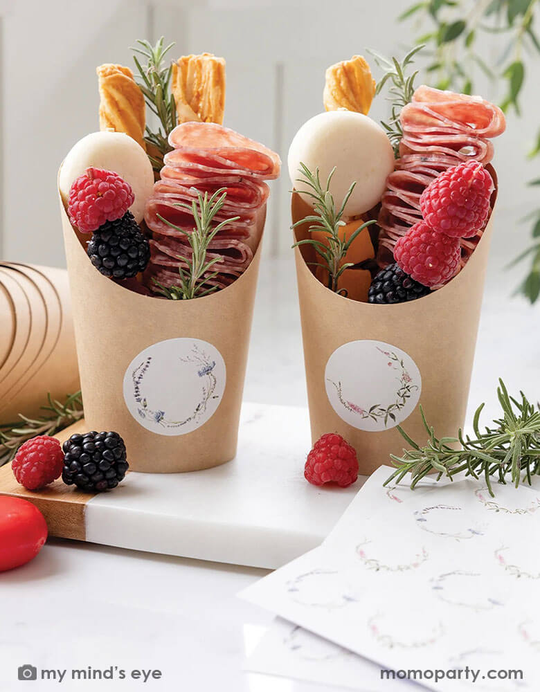 Two charcuterie cups filled with cheese, fruits and salami treats on a white marble kitchen counter. Around them are a marble cutting board with some berries on it and rosemary bunches. These charcuterie cups from Momopartys that come with floral round labels for your guests names are perfect for a spring or summer soriee, whether it's a baby shower, a bridal shower or kid's birthday parties.