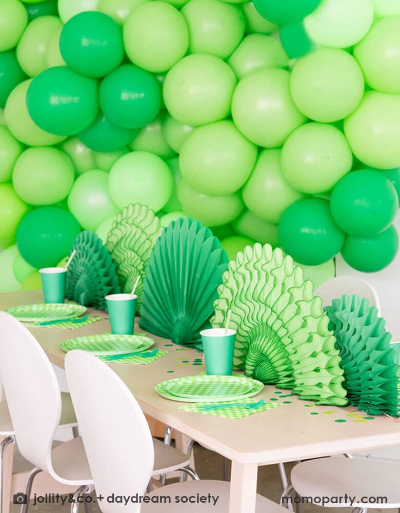 A green themed party set up featuring a giant green balloon wall in the backdrop. On the table features Momopartys's Check it! Lime Green Checkered Tableware Collection by Jollity & Co. including 10" dinner plates, 8" dessert plates, 6.5" large napkins and solid green party cups, with green paper fans as the centerpiece.