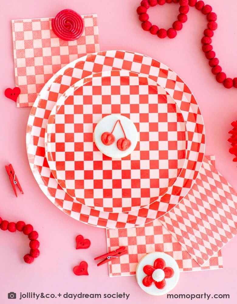 A pink and red party table featuring Momopartys's check it red checkered party collection including red checkered patterned dinner and side plates, large and small napkins by Jollity Co. With sugar cookies with red flower and cherry designs and some party favors goods and felt garland around the tableware, it makes it a modern and chic playful party table.