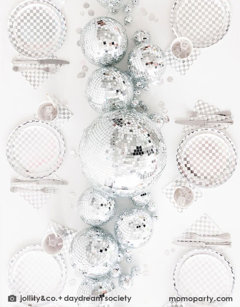 A glamorous and dazzling party table featuring Momopartys's check it! silver checkered tableware collection by Jollity & Co. including 10" dinner plates, 8" dessert plates, 6.5" large napkins, solid silver party cups and silver cutlery. In the middle of the table are a bunch of disco balls in different sizes with silver confetti scattered, what a festive party table! A perfect inspo for New Year's Eve celebration or a disco themed party!
