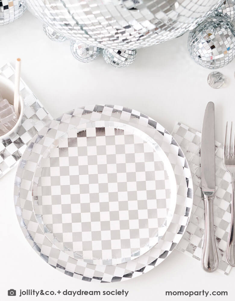 A glamorous and dazzling party table featuring Momopartys's check it! silver checkered tableware collection by Jollity & Co. including 10" dinner plates, 8" dessert plates, 6.5" large napkins, solid silver party cups and silver cutlery. In the middle of the table are a bunch of disco balls in different sizes with silver confetti scattered, what a festive party table! A perfect inspo for New Year's Eve celebration or a disco themed party!