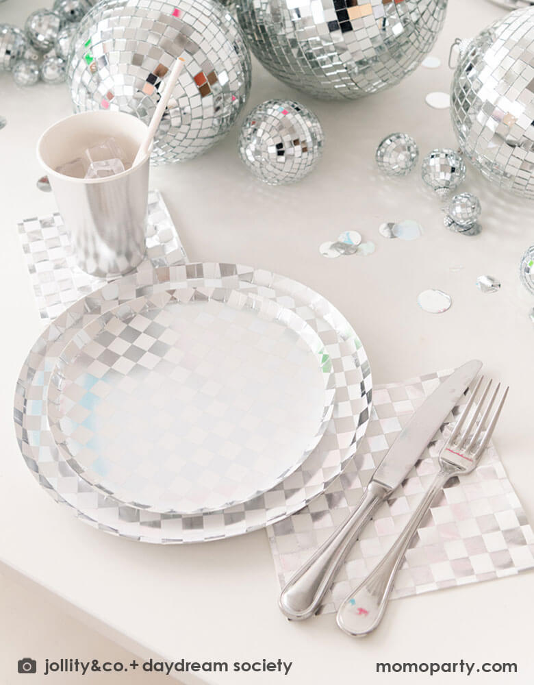 A glamorous and dazzling party table featuring Momopartys's check it! silver checkered tableware collection by Jollity & Co. including 10" dinner plates, 8" dessert plates, 6.5" large napkins, solid silver party cups and silver cutlery. In the middle of the table are a bunch of disco balls in different sizes with silver confetti scattered, what a festive party table! A perfect inspo for New Year's Eve celebration or a disco themed party!