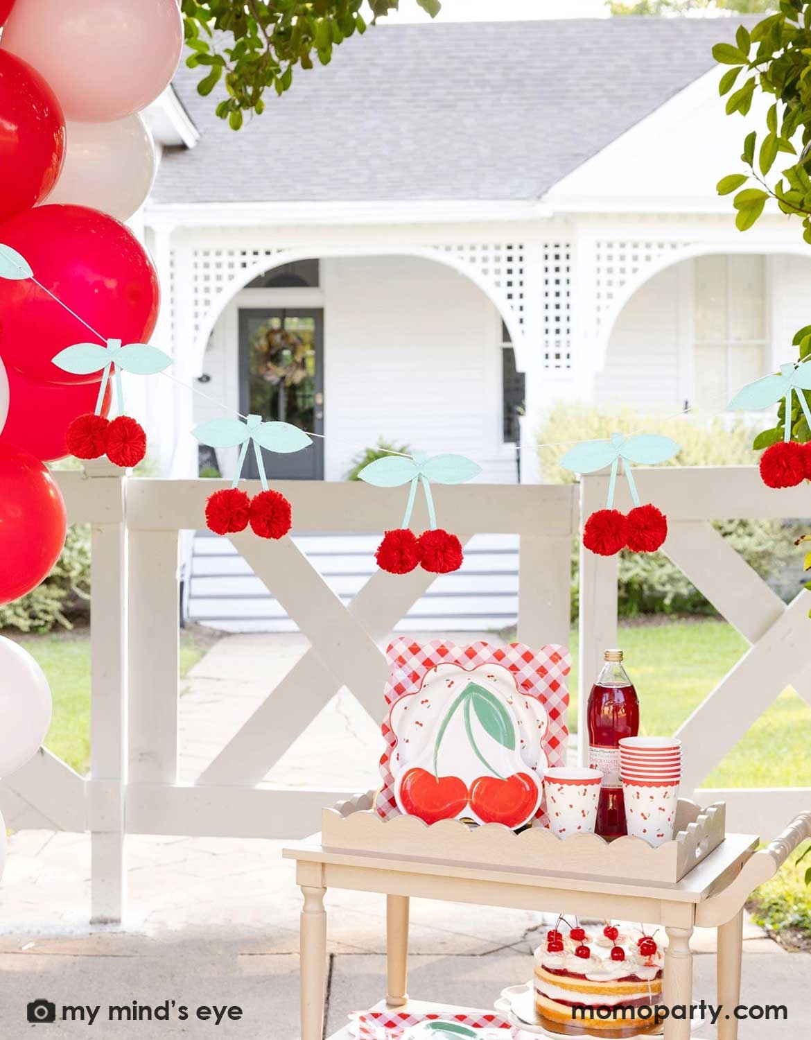 An outdoor party setting next to a red and pink balloon garland with cherry-themed decor and party tableware from Momopartys including cherry themed plates, cups, a bottle of cherry juice and a cherry cake on a wooden serving cart. Above it hung a pom pom cherry felt party banner - a simply yet adorable idea for am outdoor cherry themed party in summer.