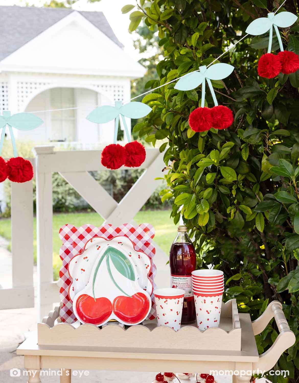 An outdoor party setting next to a tree with cherry-themed decor and party tableware from Momopartys including cherry themed plates, cups, and a bottle of cherry juice on a wooden serving cart. Above it hung a pom pom cherry felt party banner - a simply yet adorable idea for am outdoor cherry themed party in summer.