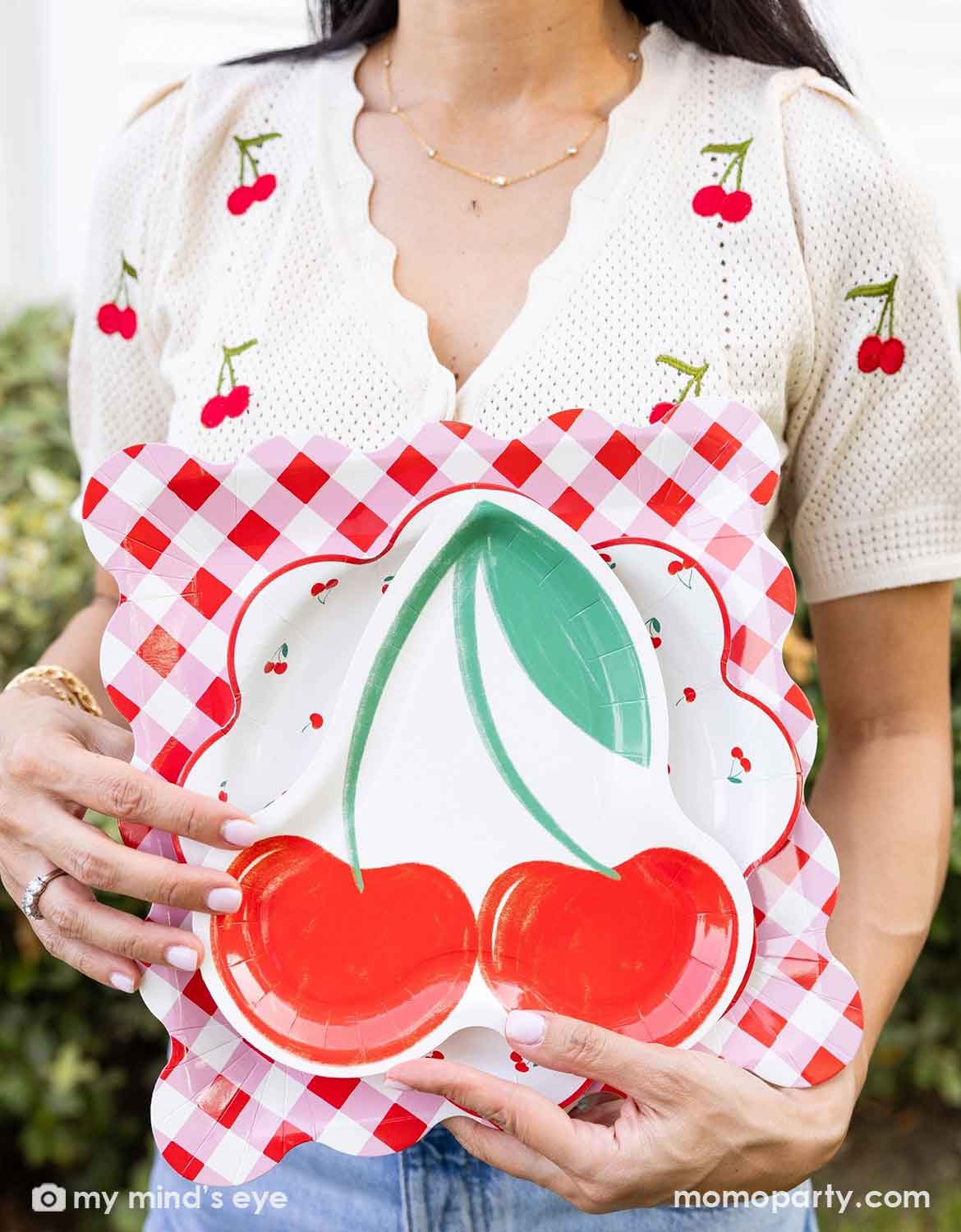 A lady in a shirt with cherry pattern on it  holding a Momopartys's cherry themed plates with cherry design and checkered pattern by Momopartys standing at an outdoor party venue.