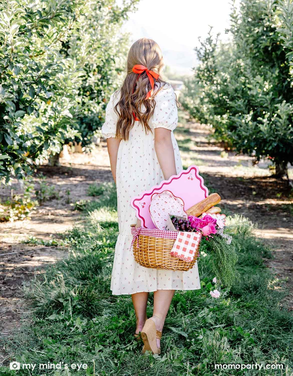 A lady in a white dress with a red cherry pattern holding a picnic basket filled with cherry themed party supplies including plates, napkins and placemats from Momopartys along with some bread and flowers walking through an orchard.