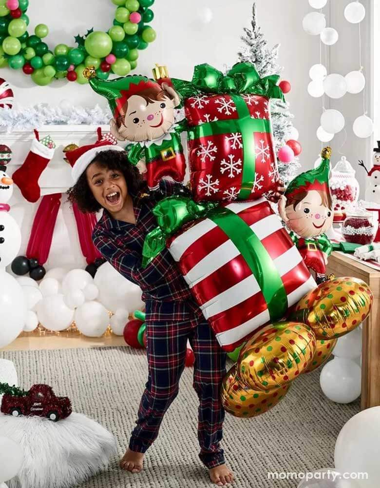 A girl in her Holiday pajama holding Momopartys's 36" x 53" Christmas Elves Airloonz balloon by Anagram Balloons. In the back it's a room decorated with Christmas balloons including a wreath, a snowman and some white balloon garland on the floor resembles snow. In the mantle there are stockings with gifts inside, makes it a perfect inspo for a fun Holiday gathering set up.