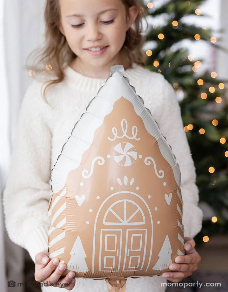 A girl in her cream white sweater holding Momopartys's 13.5" x 20" gingerbread house shaped foil balloon standing next to a big window. Behind her is a big Christmas tree lit up with small twinkling lights,