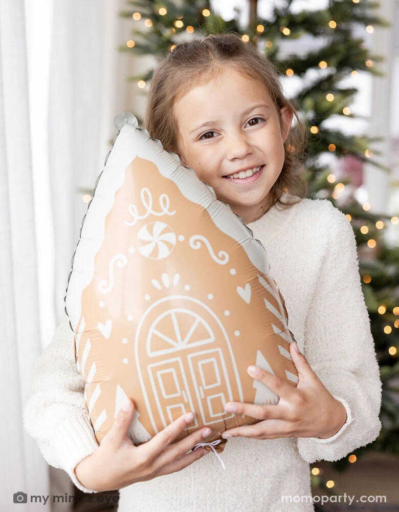 A girl in her cream white sweater holding Momopartys's 13.5" x 20" gingerbread house shaped foil balloon standing next to a big window. Behind her is a big Christmas tree lit up with small twinkling lights,