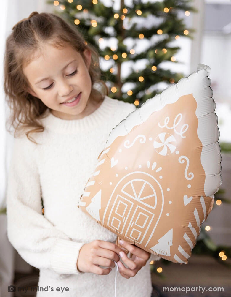 A girl in her cream white sweater holding Momopartys's 13.5" x 20" gingerbread house shaped foil balloon standing next to a big window. Behind her is a big Christmas tree lit up with small twinkling lights,