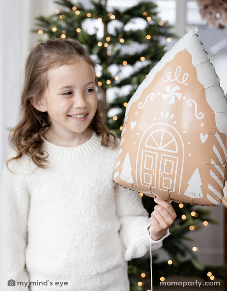 A girl in her cream white sweater holding Momopartys's 13.5" x 20" gingerbread house shaped foil balloon standing next to a big window. Behind her is a big Christmas tree lit up with small twinkling lights,