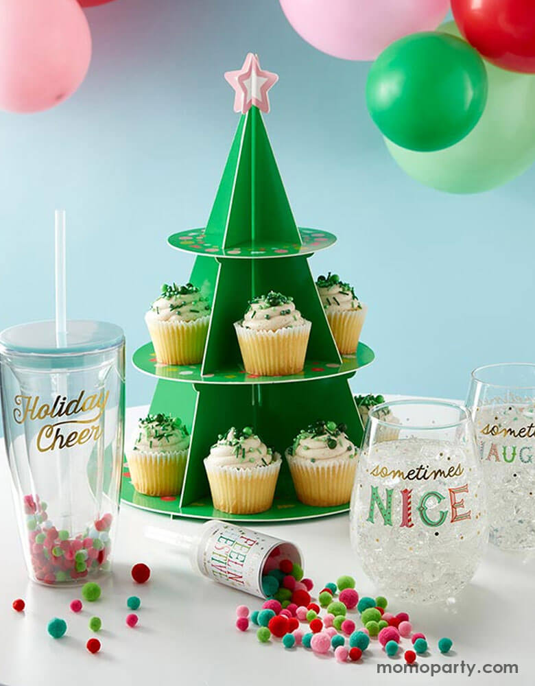 A festive Holiday party table features Momopartys's Christmas tree shaped cupcake stand with cupcakes with green sprinkles on them. On the table there are some tumblers and cups and a confetti cannon filled with mini pom poms in Christmas themed color, i.e red, green, and pink. Behind the table are some festive party balloons in red, pink, green and mint making this a perfect inspo for a festive Christmas party table ideas.