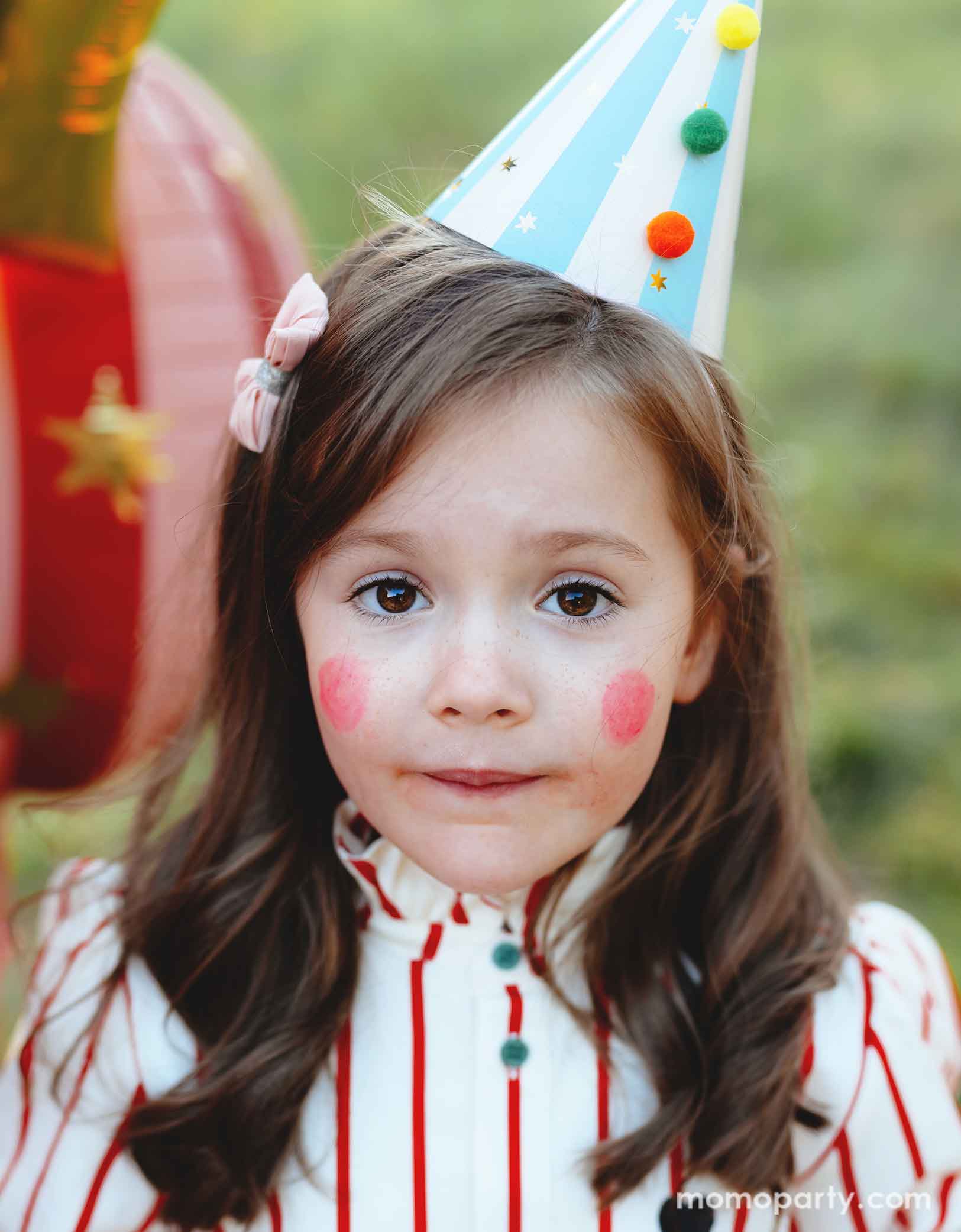 A girl dressed in vintage circus inspired outfits and make up wearing Momopartys's light blue striped Birthday Party Hat with Pompoms, in a festive carnival themed event. In her back there are some fun circus themed foil party balloons floating. 