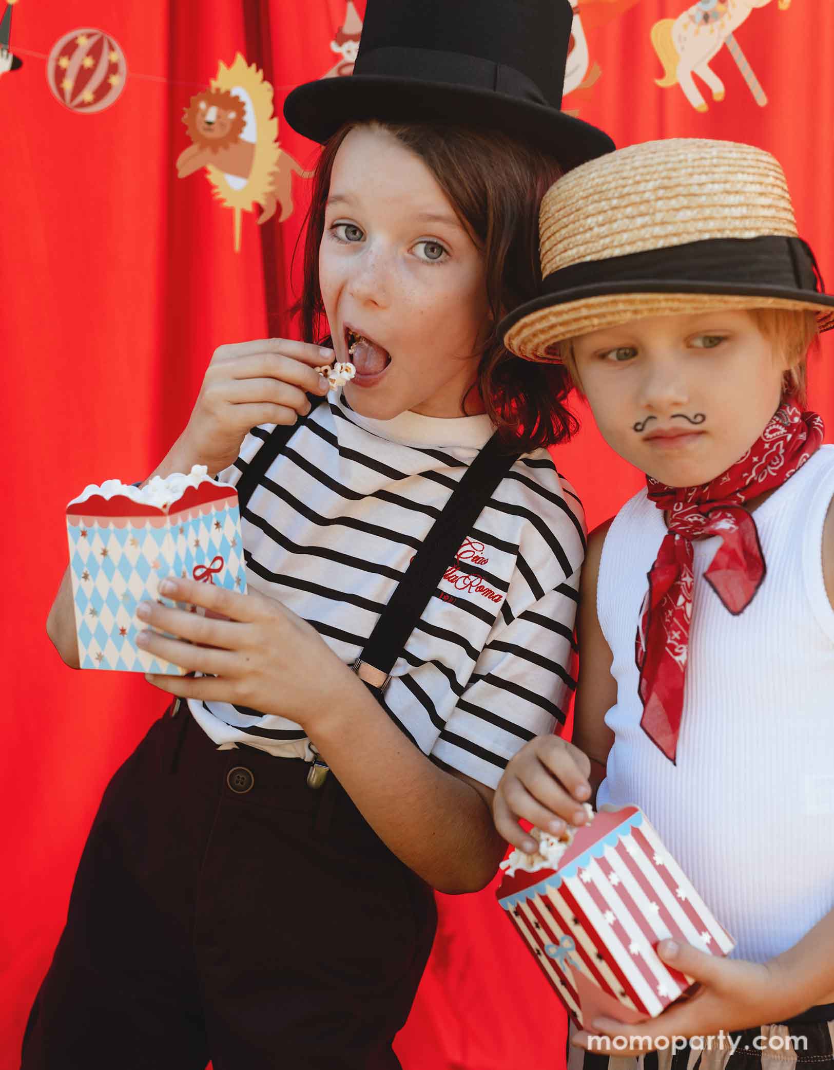 Two kids dressed in the vintage circus outfits and makeup holding Momopartys's Circus Striped Treat Boxes filled with popcorn in their hands in a festive carnival themed event. In their back is a red stage curtain as a party backdrop decorated with circus themed birthday banner with circus animal party pennants. 