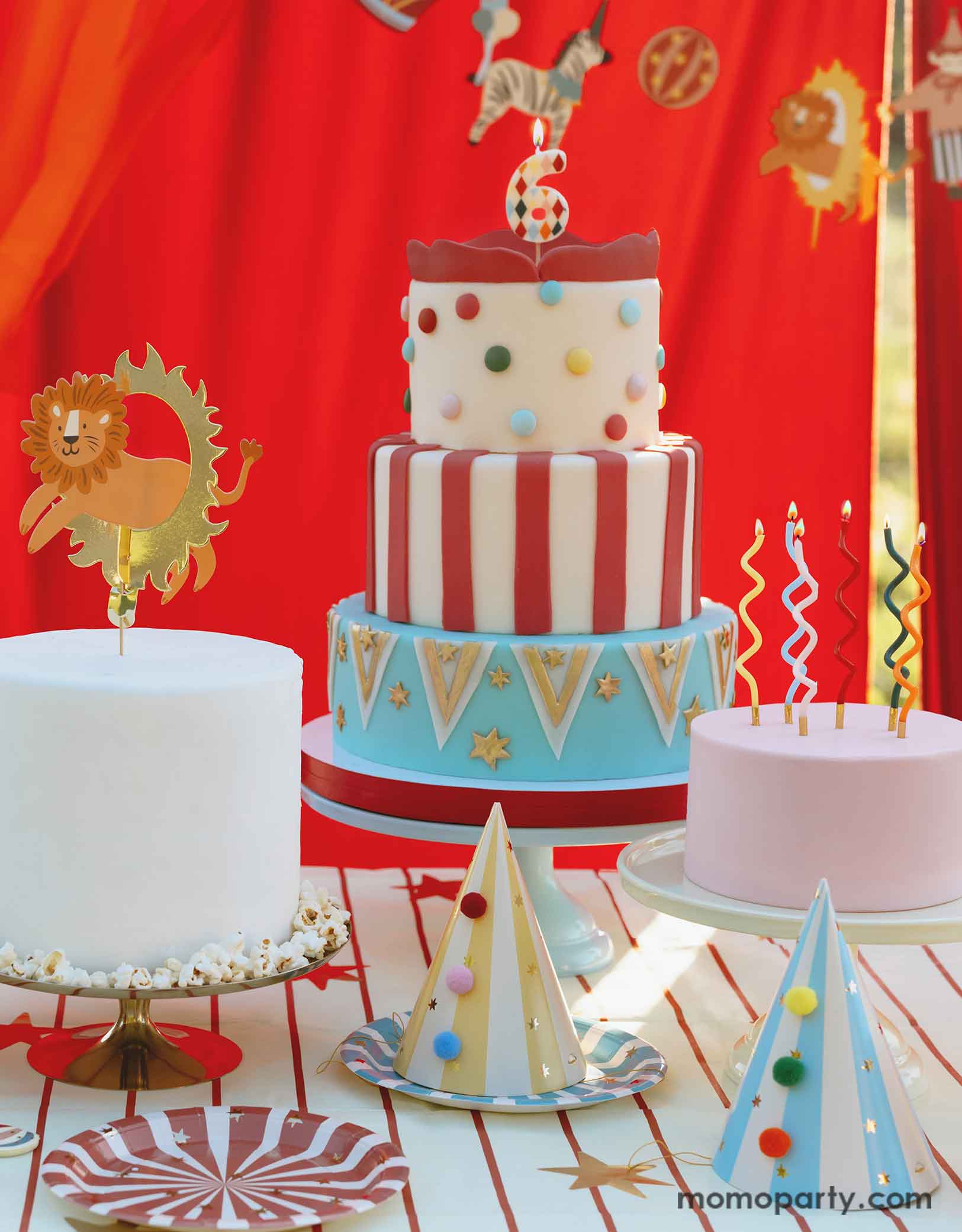 A circus party table featuring multiple carnival themed birthday cakes topped with fun candles and a lion jumping the loop cake topper. On the table there are  Momopartys's circus themed party tableware including colorful striped paper plates in 6 different colors and striped party hats with pom poms. In the back there's a red curtain backdrop adorned with circus happy birthday banner, making this great table setting idea for a fun and festive kid's carnival/circus themed birthday celebration.