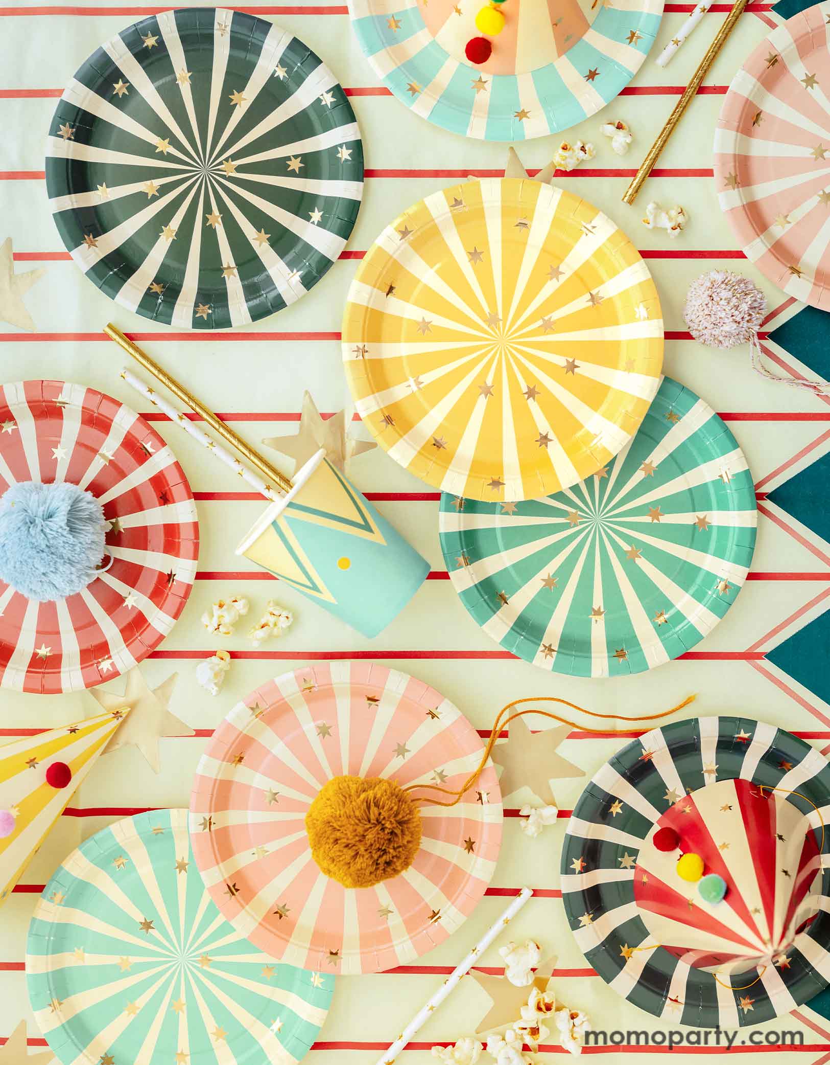 A carnival/circus themed part table featuring Momopartys's circus themed party tableware including colorful striped paper plates in 6 different colors, matching party cups, and striped party hats with pom poms, with gold foil straws and popcorns on a circus themed striped table cover - making this great table setting idea for a fun and festive kid's carnival themed birthday celebration!