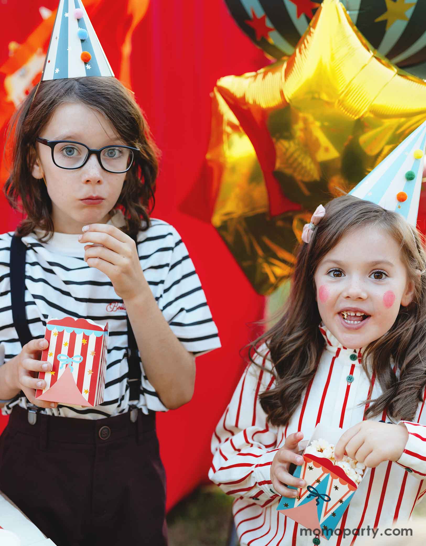 Two kids dressed in the vintage circus outfits and makeup holding Momopartys's Circus Striped Treat Boxes filled with popcorn in their hands, wearing party hats with colorful pom poms in a festive carnival themed event. In their back is a red stage curtain as a party backdrop decorated with circus themed birthday banner with circus animal party pennants and some festive large carnival themed foil balloons. 