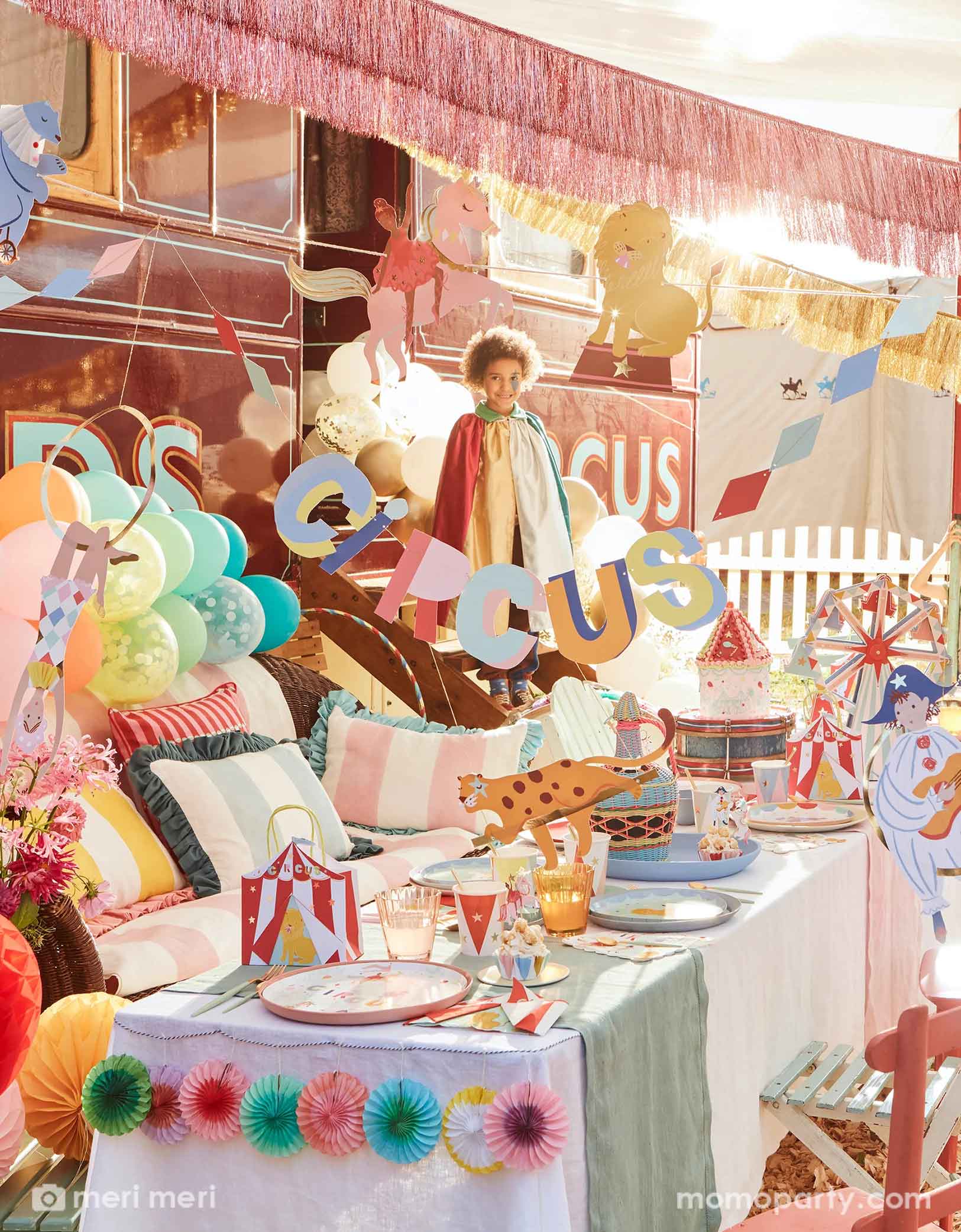 A festive circus themed party featuring Momopartys's circus themed party supplies including a table filled with circus themed party tablewares including plates, napkins, party cups, party bags and ferry's wheel cupcake stand. There's the circus party garland featuring circus animals with honeycomb decorations hung above the party table. Above the party garland, there are pink and gold fringe garlands. Around the table there are some kids dressed up in circus inspired costumes, making this a playful scene.  