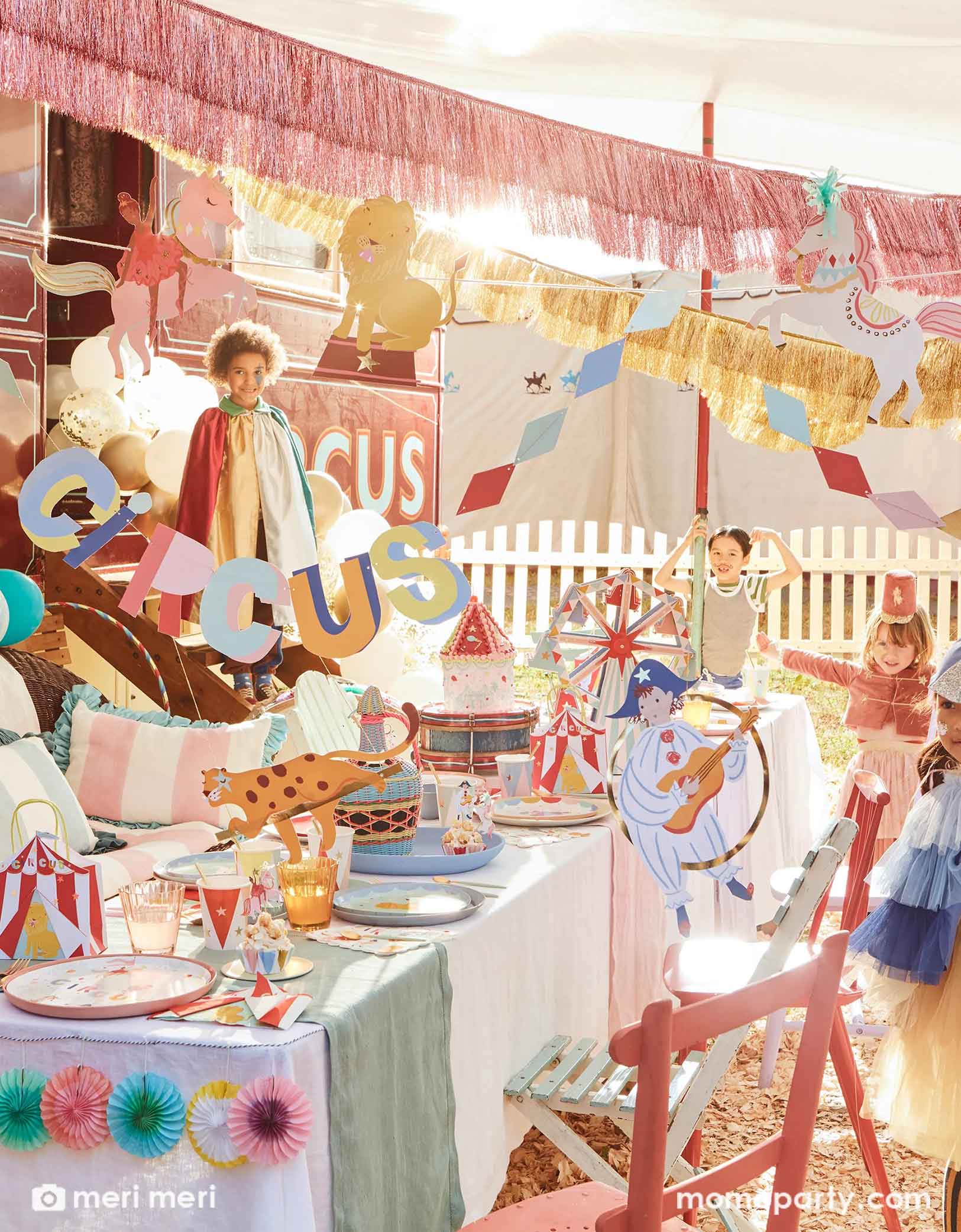A festive circus themed party featuring Momopartys's circus themed party supplies including a table filled with circus themed party tablewares including plates, napkins, party cups, party bags and ferry's wheel cupcake stand. There's the circus party garland featuring circus animals with honeycomb decorations hung above the party table. Above the party garland, there are pink and gold fringe garlands. Around the table there are some kids dressed up in circus inspired costumes, making this a playful scene.  