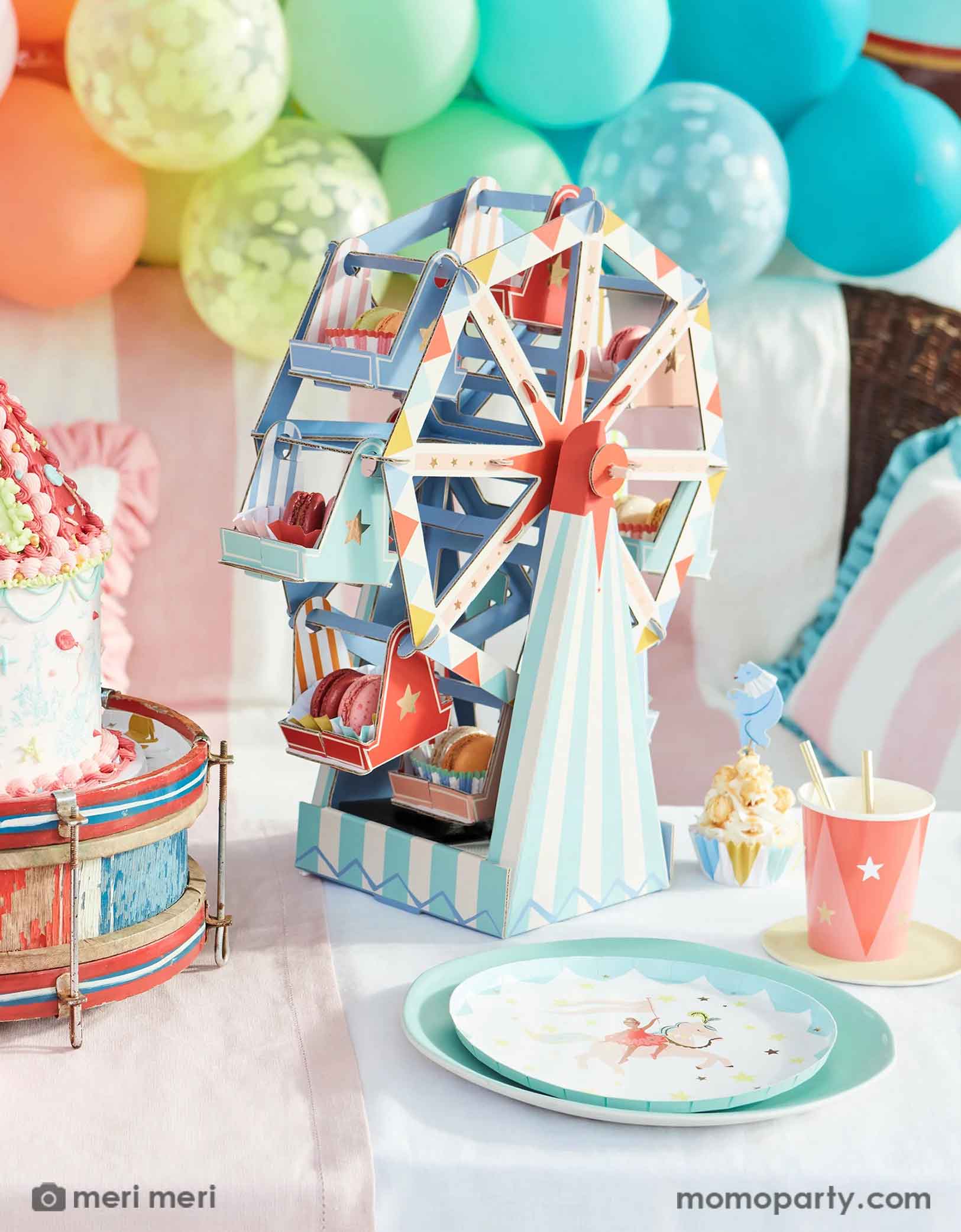 A festive carnival, circus themed party featuring Momopartys's circus themed party supplies and decorations by Meri Meri including the circus side plates, party cups, cupcake kit, ferry's wheel cupcake stand and a rainbow colored balloon garland in the back, with a circus tent shaped birthday cake on a vintage drum and pastel striped table cloth, this makes a great inspiration for a playful and fun circus party decoration idea.