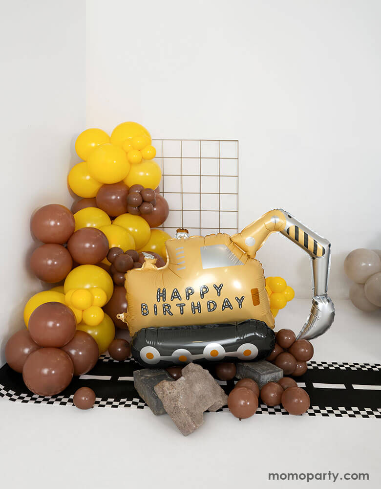 A construction themed birthday party balloon set up featuring Momopartys's digger shaped happy birthday foil balloon next to a balloon garland in brown and yellow. On the floor there's a road table runner. Making this a fun decoration for kid's construction themed birthday celebration!