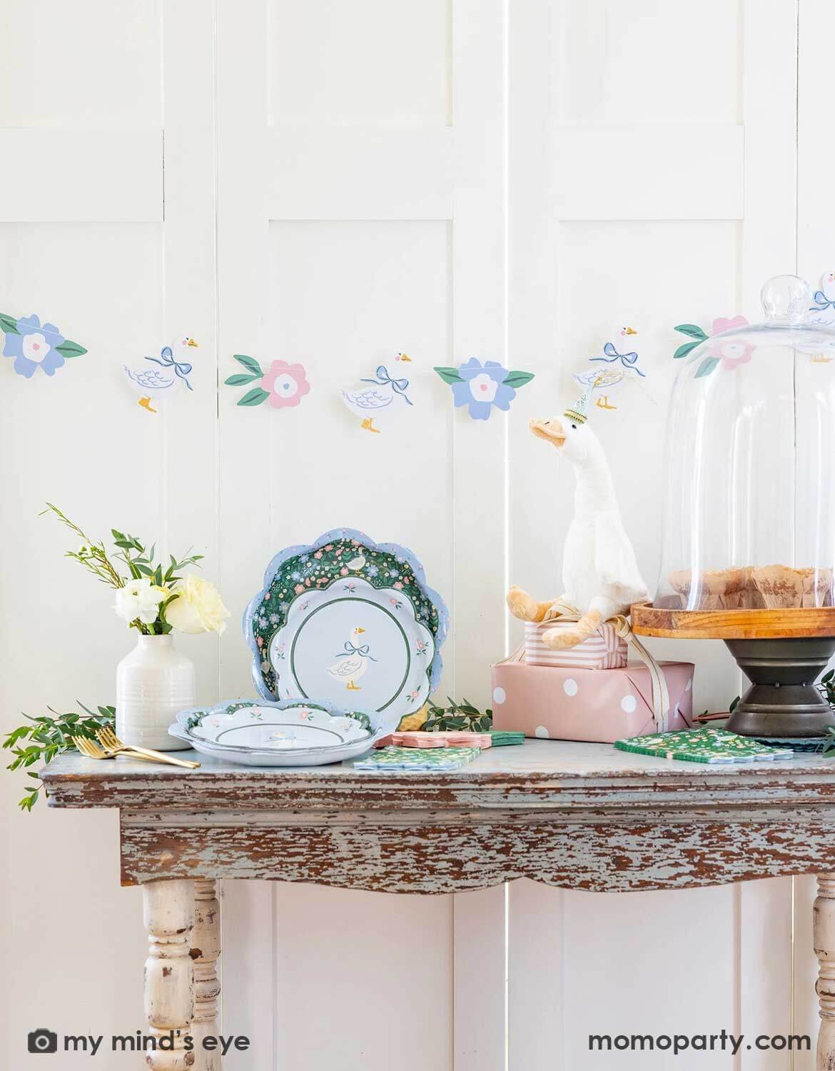 A rustic vintage style table setting featuring various goose themed party supplies and decorations from Momopartys including goose plates, napkins and party banner with floral design on a white wall background. On the table there are also some goose decoration and flowers, making this a perfect setting for kid's Silly Goose themed birthday party, a baby shower or an Easter celebration.