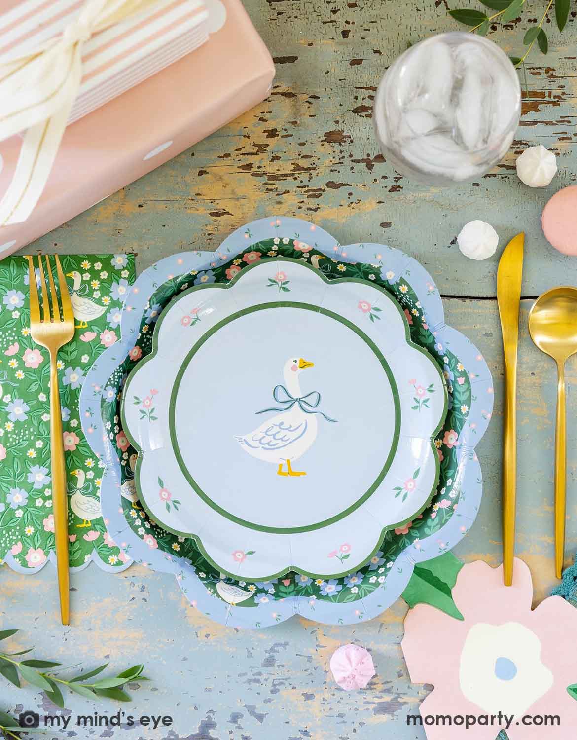 A rustic cottage inspired tablescape featuring Momopartys's goose paper plates and floral napkins with gold cutlery and flowers on a rustic blue table. A perfect table setting idea for a kid's "One Silly Goose" themed 1st birthday party, a boy baby shower or an Easter celebration. 