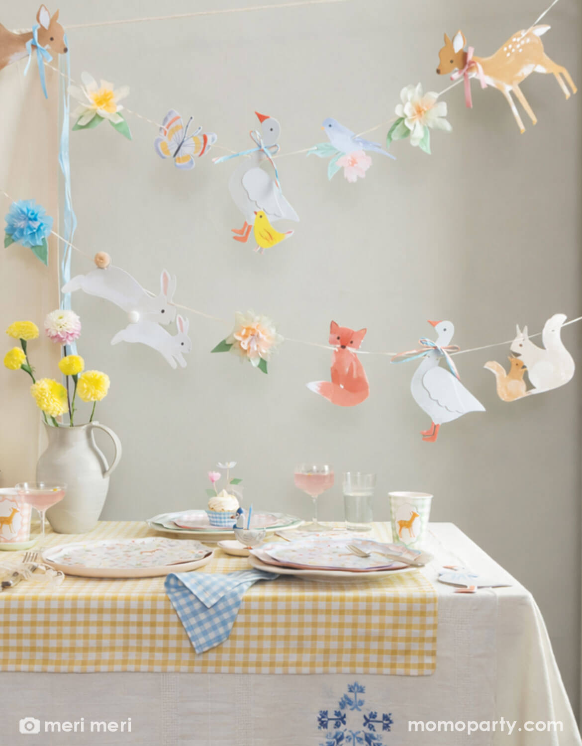 A rustic cottage themed table setting with little woodland animals of deer, goose, bunny, fox, chicks in tablewares and decorations from Momopartys - including party cups, matching plates and napkins and an adorable little animals party garland hung above the table covered with a soft gingham tablecloth. With spring florals in soft yellow and pink on the table, this table is perfect for a baby shower, an Easter celebration or young kid's storybook rustic cottage inspired birthdays.