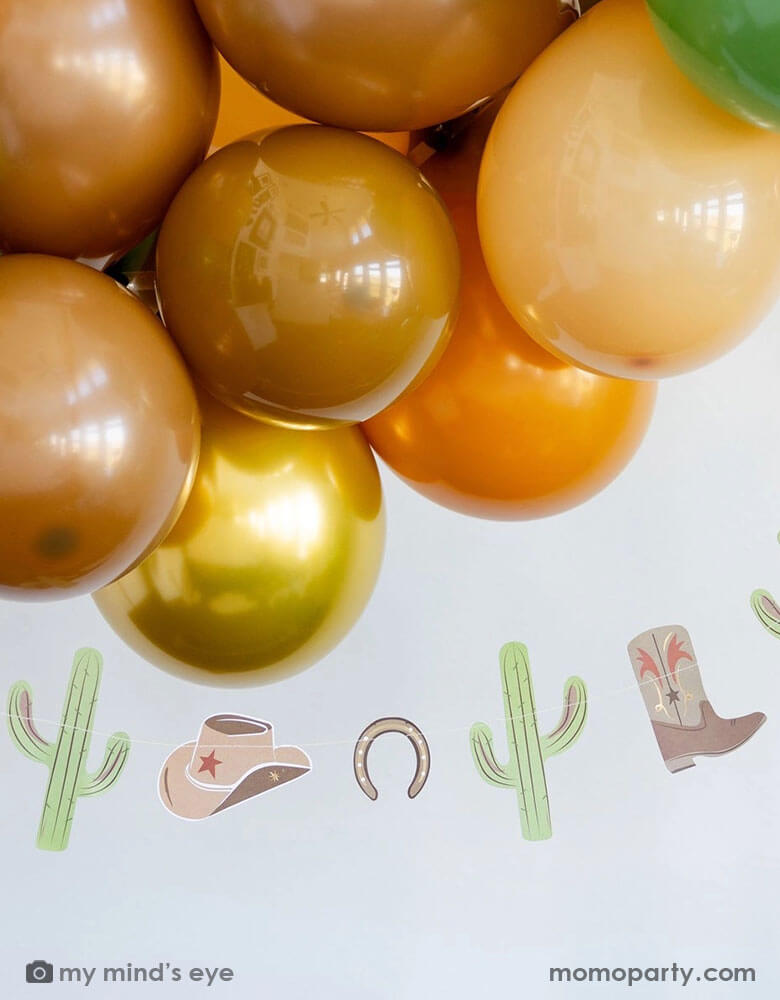A western cowboy themed party decoration featuring Momopartys's cowboy icon party banner by My Mind's Eye and a balloon garland with green, brown, and gold colors - a perfect yet simple decoration idea for a Western themed celebration, be it a First Rodeo first birthday party for boys or Wild Wild West bash!