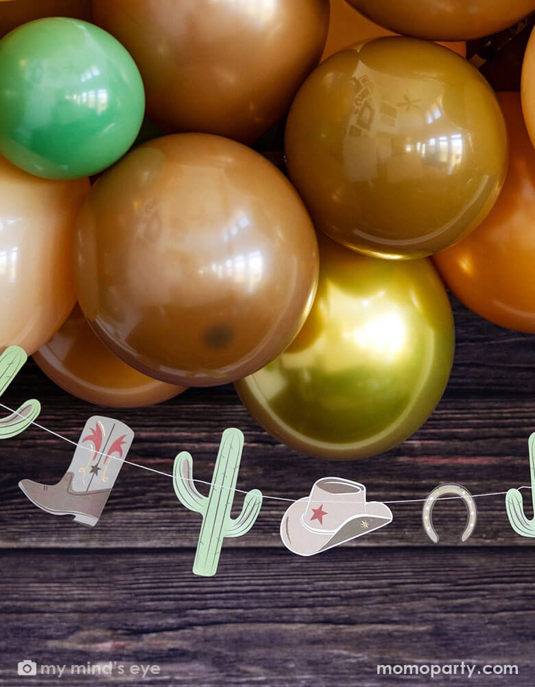 A western cowboy themed party decoration featuring Momopartys's cowboy icon party banner by My Mind's Eye and a balloon garland with green, brown, and gold colors, with the rustic wooden background, this makes a perfect inspiration for a Western themed celebration, be it a First Rodeo first birthday party for boys or Wild Wild West bash!
