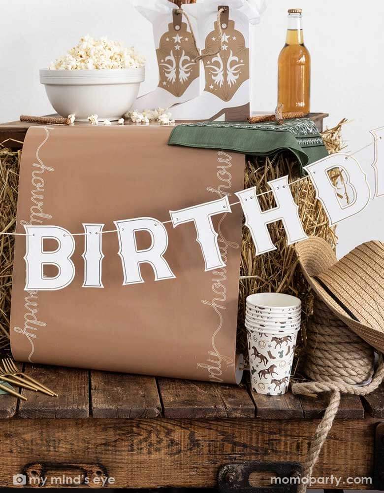 A cowboy themed party table featuring Momopartys's cowboy themed party supplies including cowboy patterned paper cups, birthday banner, table runner and cowboy boot shaped treat boxes. On the table there's a cowboy hat, some straw bales, and a green bandana as decoration, with popcorn, treats and soda bottle, it's perfect for a Western rodeo themed celebration, be a First Rodeo boy's first birthday party or a fun cowboy bash!