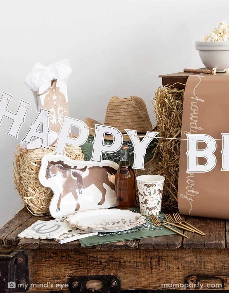 A cowboy themed party table featuring Momopartys's cowboy themed party supplies including horse shaped plates, cowboy patterned cups, birthday banner, napkins, table runner and cowboy boot shaped treat boxes. On the table there's a cowboy hat, straw bale, and a green bandana as decoration, perfect for a Western rodeo themed celebration, be a First Rodeo boy's first birthday party or a fun cowboy bash!