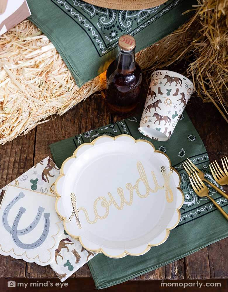 An overhead shot of a cowboy themed party tablescape featuring Momopartys's cowboy themed party supplies including howdy paper plates, cowboy patterned cups, and napkins. On the table there's a straw bale, and a green bandana as decoration, and a soda bottle, perfect for a Western rodeo themed celebration, be a First Rodeo boy's first birthday party or a fun cowboy bash!