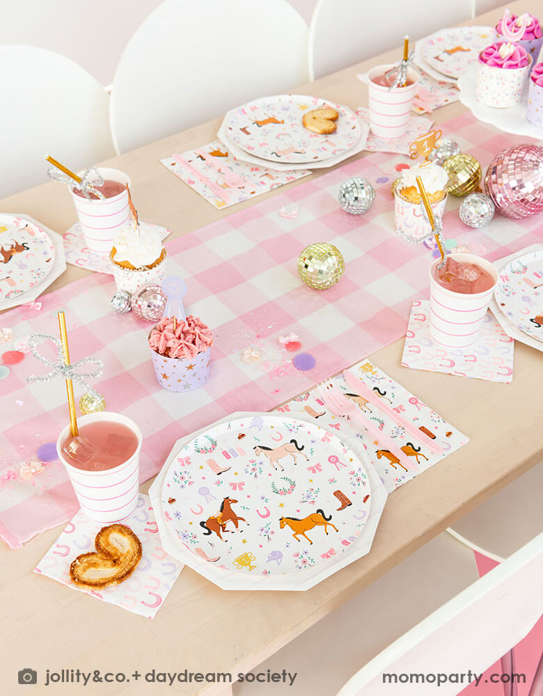 A girly cowgirl rodeo pony themed party table featuring Momopartys's cowgirl pony themed party plates, napkins, horseshoe napkins, pink striped party cups and a pink gingham table runner. Around the tableware there are disco ball decorations and a cake stand with cupcakes with pink icing, making this an adorable inspiration for girl's First Rodeo first birthday celebration or an adorable cowgirl theme party for pony-loving girls!