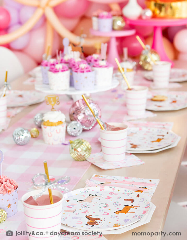 A close up of a girly cowgirl rodeo pony themed party table featuring Momopartys's cowgirl pony themed party plates, napkins, horseshoe napkins, pink striped party cups and a pink gingham table runner. Around the tableware there are disco ball decorations and a cake stand with cupcakes with pink icing. Behind the table there is a balloon wall with pink balloons, making this an adorable inspiration for girl's First Rodeo first birthday celebration or an adorable cowgirl theme party for pony-loving girls!