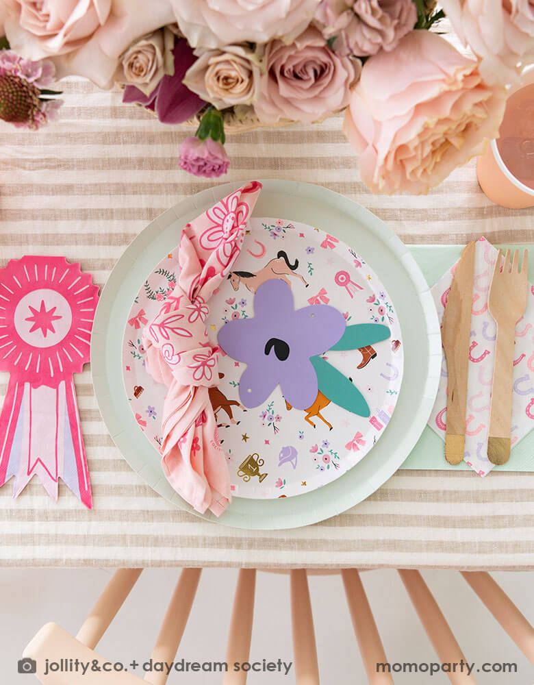 An overhead shot of a girly pony themed party tablescape featuring Momopartys's cowgirl pony themed party plate, horseshoe napkin, pink ribbon award shaped napkin and gold dipped wooden cutlery, with a pink bandana, all on a mocha brown striped tablecloth. There's some beautiful rose bouquet as the centerpiece, making this an adorable inspiration for girl's pony horse themed birthday celebration.