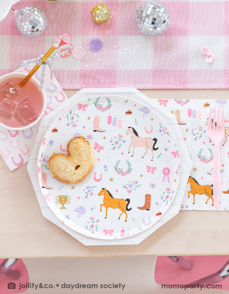 A close up of a girly cowgirl rodeo Western themed party tablescape featuring Momopartys's cowgirl pony themed party plate, napkin, horseshoe napkin, pink striped party cups and a pink gingham table runner. Around the tableware there are some lilac round confetti spread-out and a small pink disco ball decoration, making this an adorable inspiration for girl's First Rodeo first birthday celebration or an adorable cowgirl theme party for pony-loving girls!