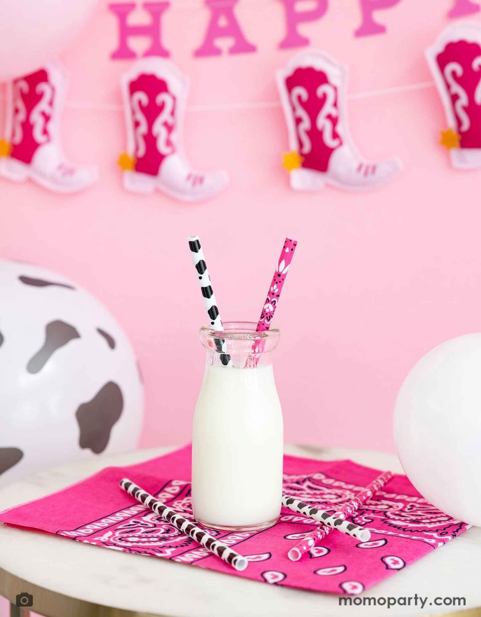 A fun cowgirl party table featuring a hot pink bandana with a bottle of milk with Momopartys's cowgirl party reusable straws in pink and cow print design. In the back of the party table are some cow print balloons and a cowgirl boot Happy birthday party banner hung on a pink wall. Making this a fun yet simple decorating idea for a cowgirl disco celebration - whether it's a "First Rodeo" girl's first birthday party or a fun cowgirl bachelorette party.